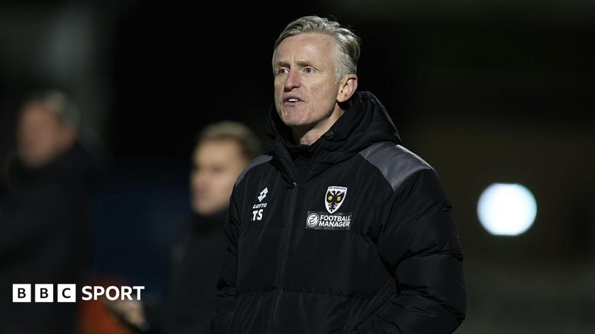 AFC Wimbledon assistant manager Terry Skiverton looks on from the touchline wearing a black coat